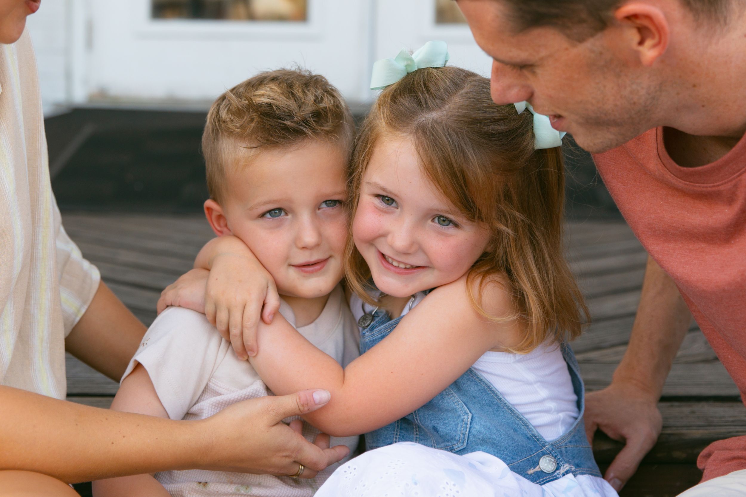 Two children hugging and smiling on an outdoor bench while adults look on.