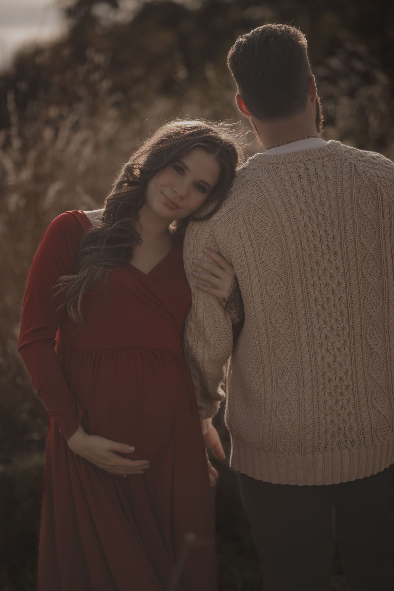 An expectant couple embraces in warm lighting, with woman in red dress and man in cream sweater.