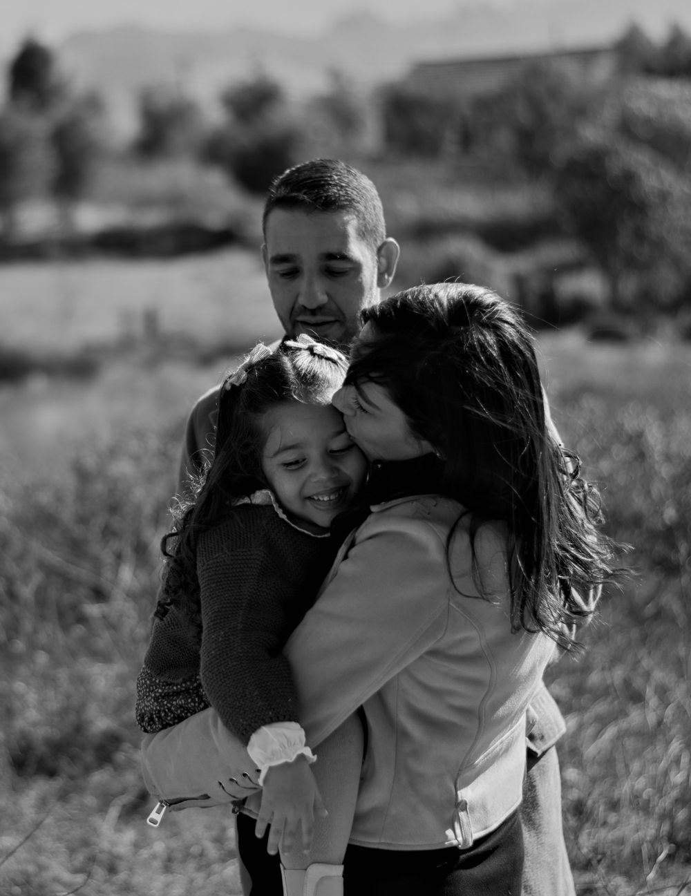Abraço entre mãe, pai e filha durante uma sessão fotográfica de família ao ar livre.