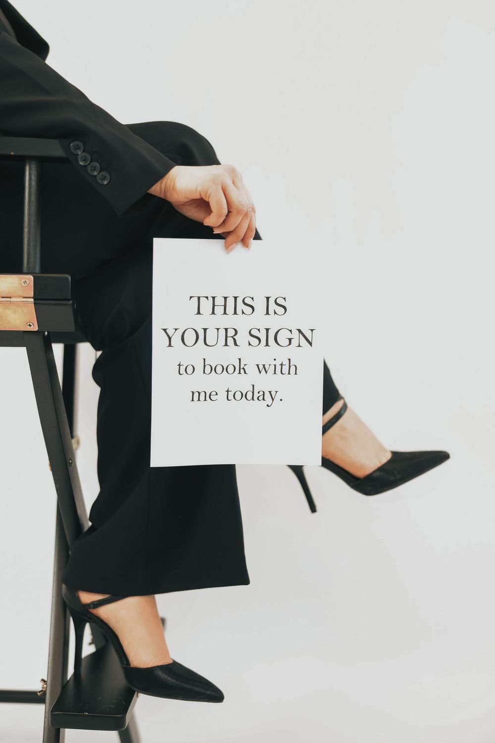 A person in black suit and heels holds a sign reading This Is Your Sign to book with me today.