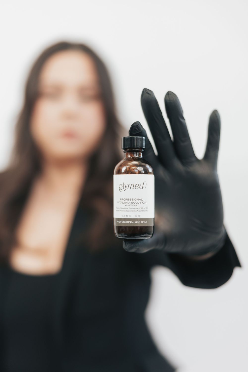 Woman in black gloves holding a GlyMed professional vitamin solution serum bottle against white background.