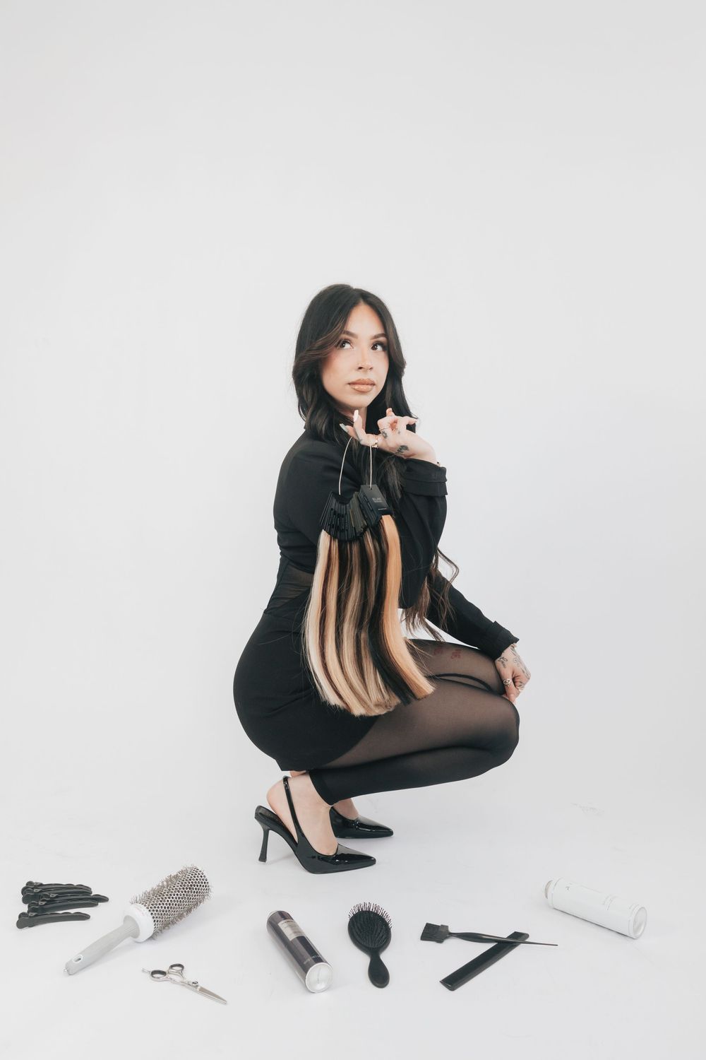 Female hairstylist in black dress crouching with hair extensions, surrounded by professional hair tools on white background.