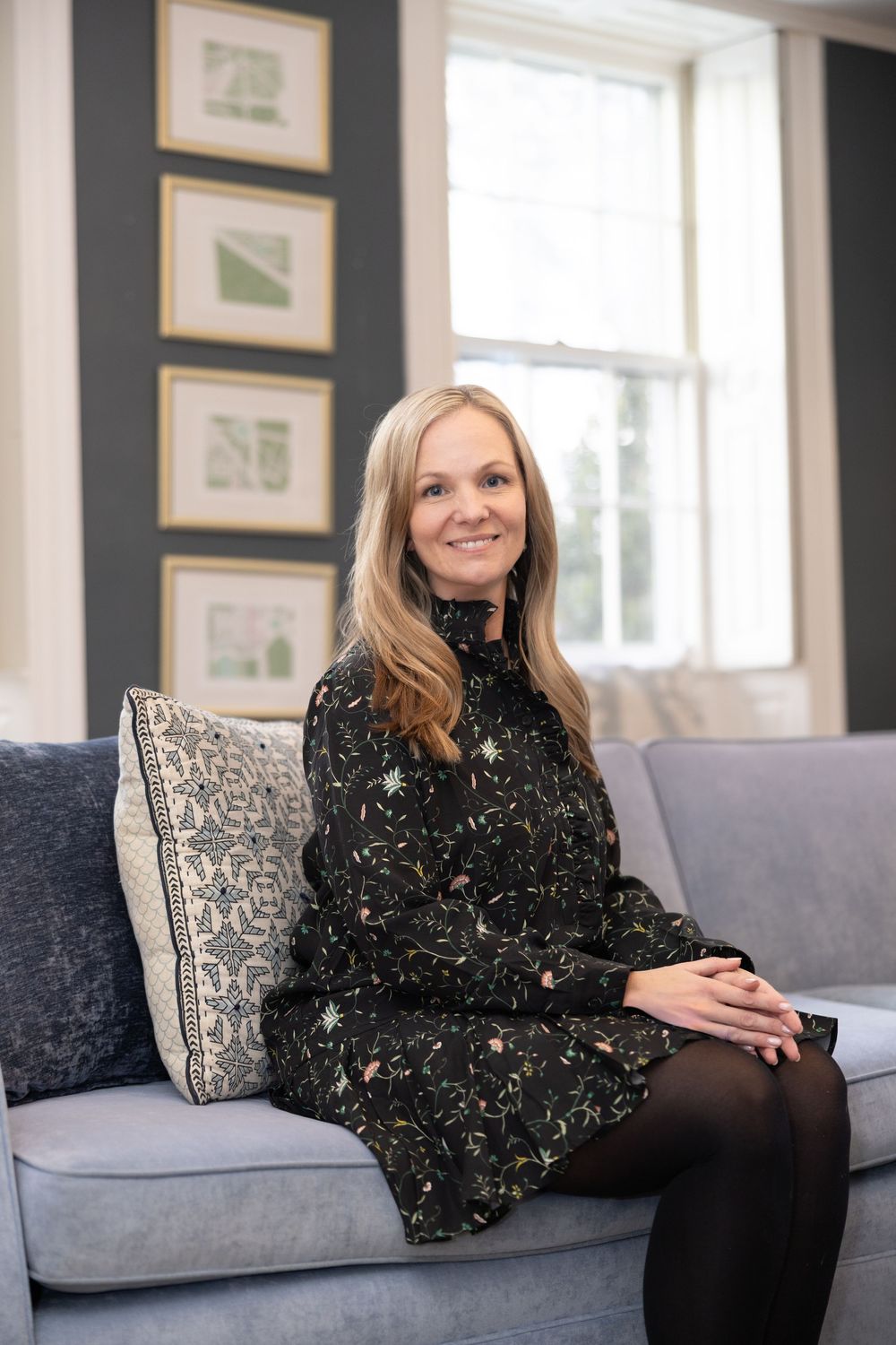 A woman in a black floral dress sits comfortably on a gray couch in a room with framed artwork on dark walls.
