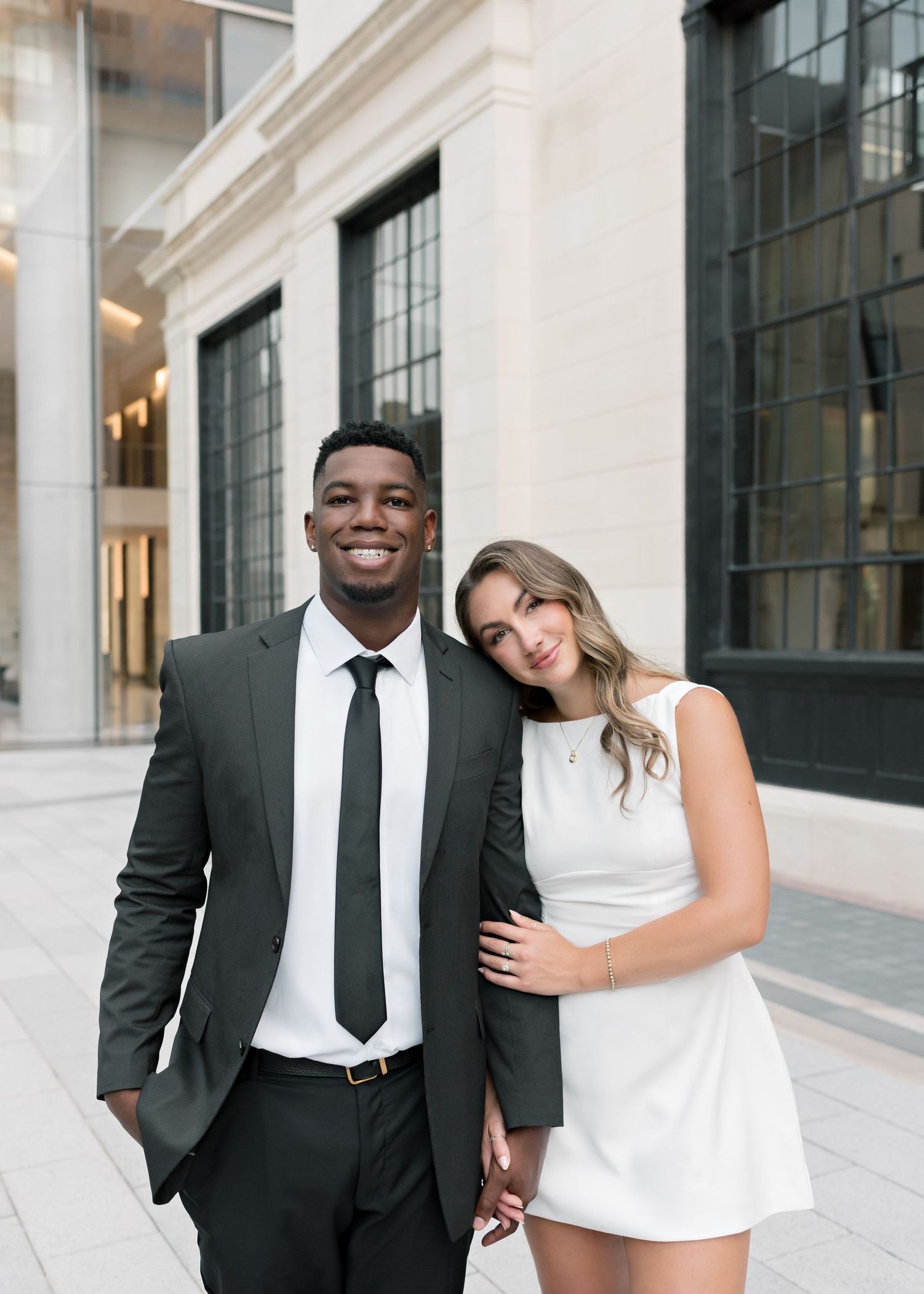 A happy couple stands together in formal attire outside a modern building.
