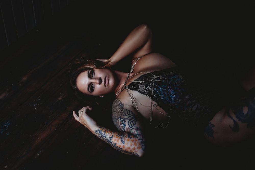 A moody boudoir photo features a figure in dark lighting wearing lace lingerie and displaying tattoo artwork.