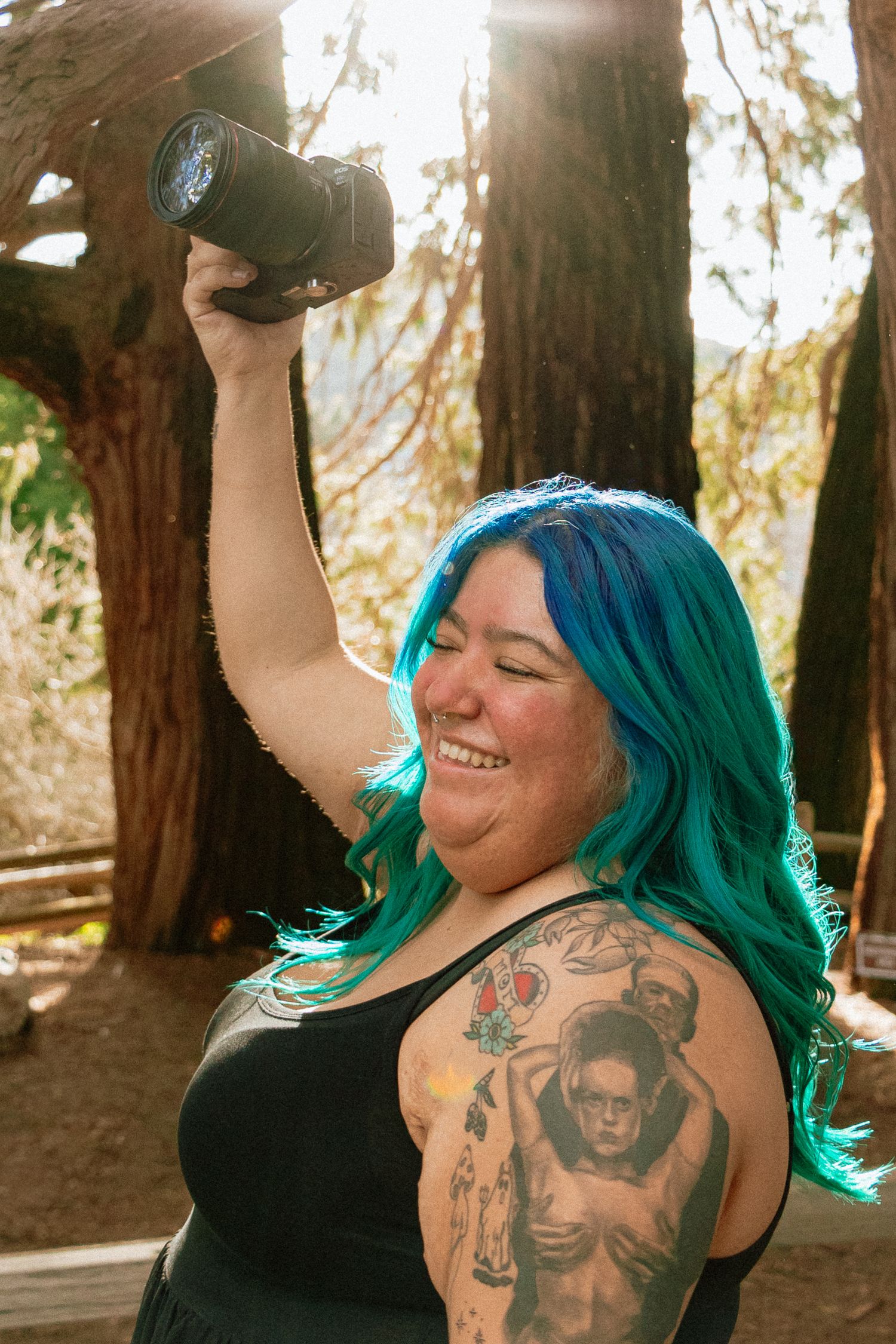 A Latina woman photographer with vibrant teal hair and tattoos holds up their camera in a sunlit forest setting.