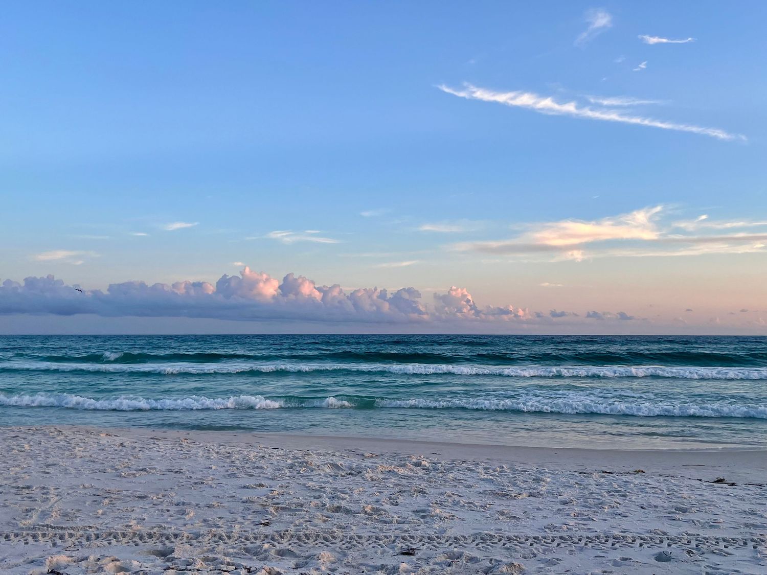 Turquoise waves roll onto a white sandy beach under a blue sky with wispy clouds at sunset.
