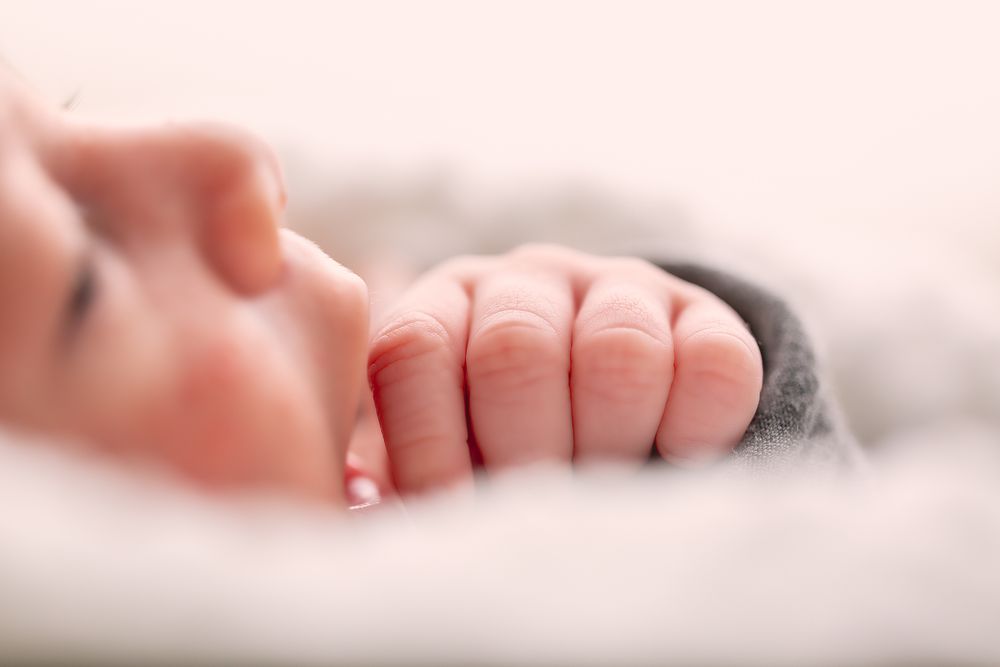 Close-up of a sleeping newborn baby's tiny clenched fist resting on a soft white blanket.