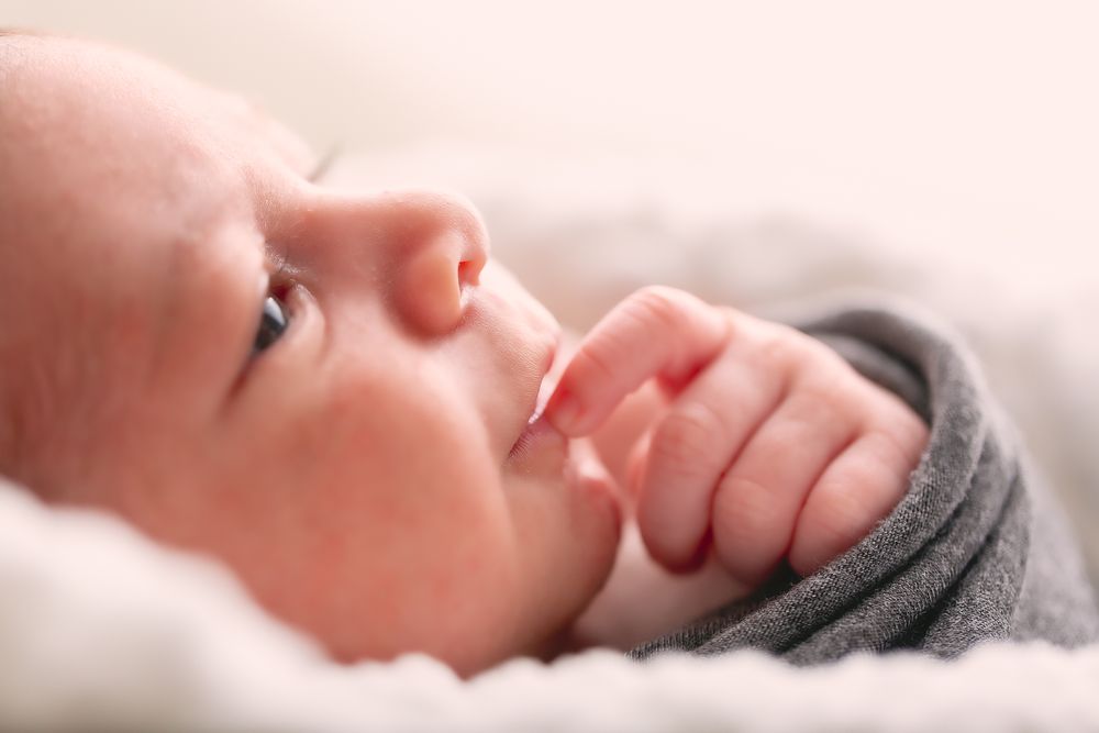 Close-up of a peaceful newborn baby lying on a white blanket with fingers near mouth.
