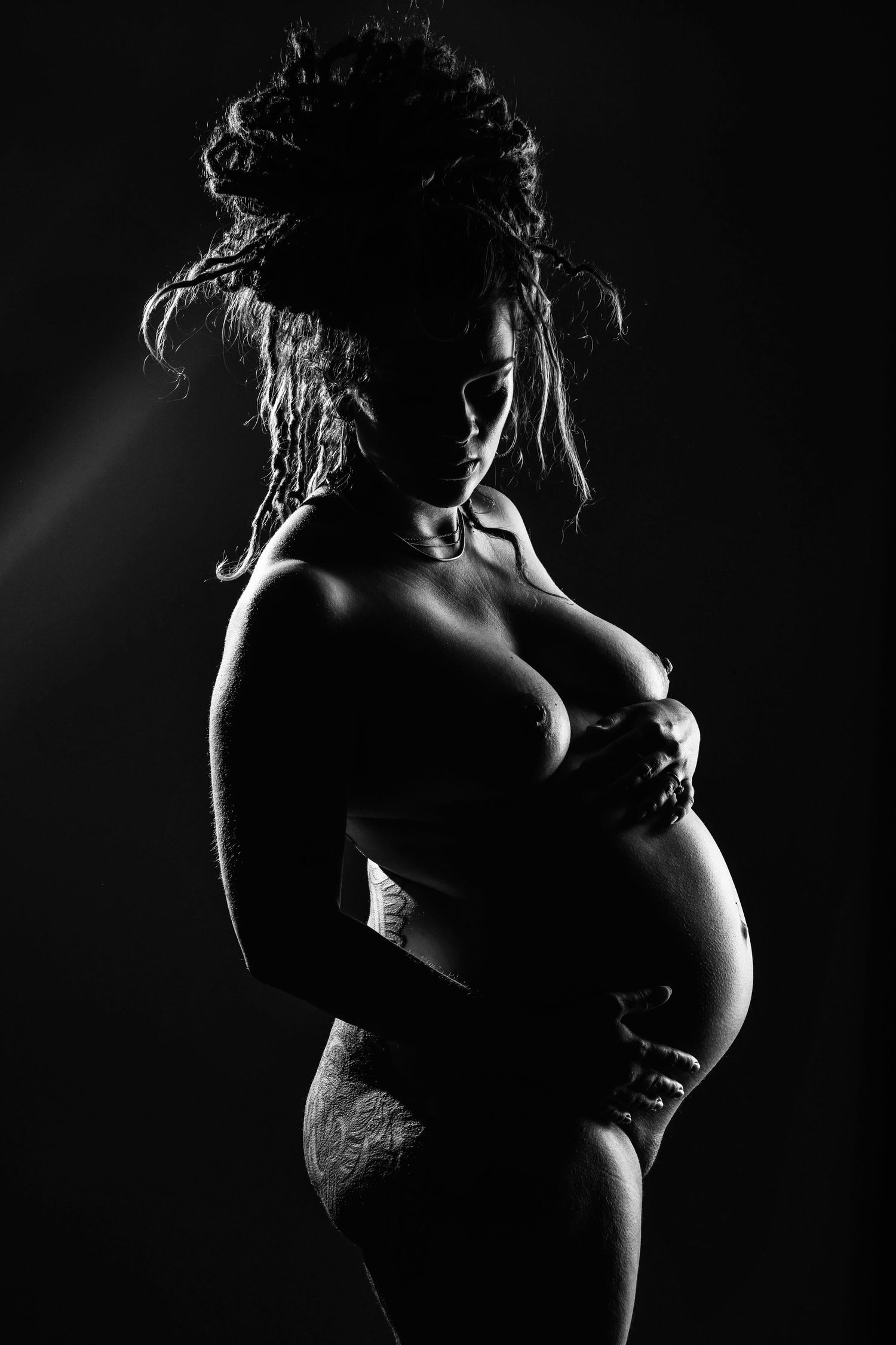 Zwart-wit silhouet van een zwangere vrouw met dreadlocks, dramatisch belicht tegen een donkere achtergrond