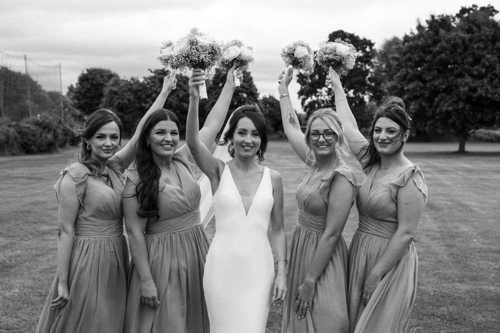 Bridesmaids in matching dresses hold bouquets overhead in outdoor setting.