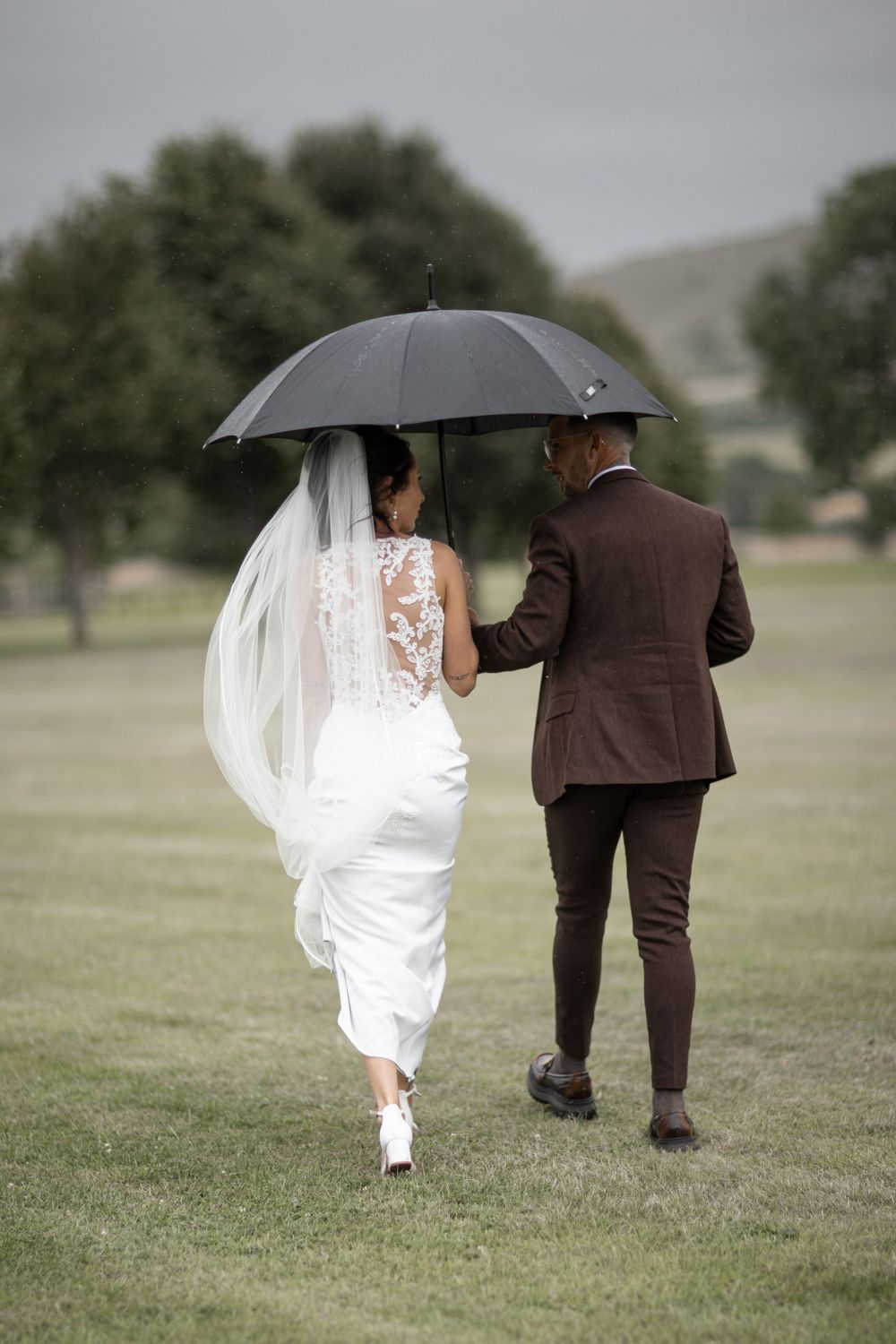 Couple walking across a field sharing an umbrella on their rainy wedding day.