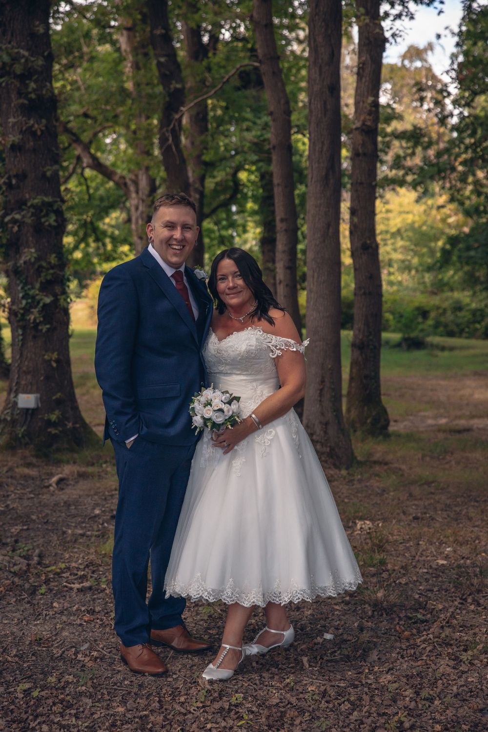 Wedding couple poses in wooded area with bride in tea-length dress.