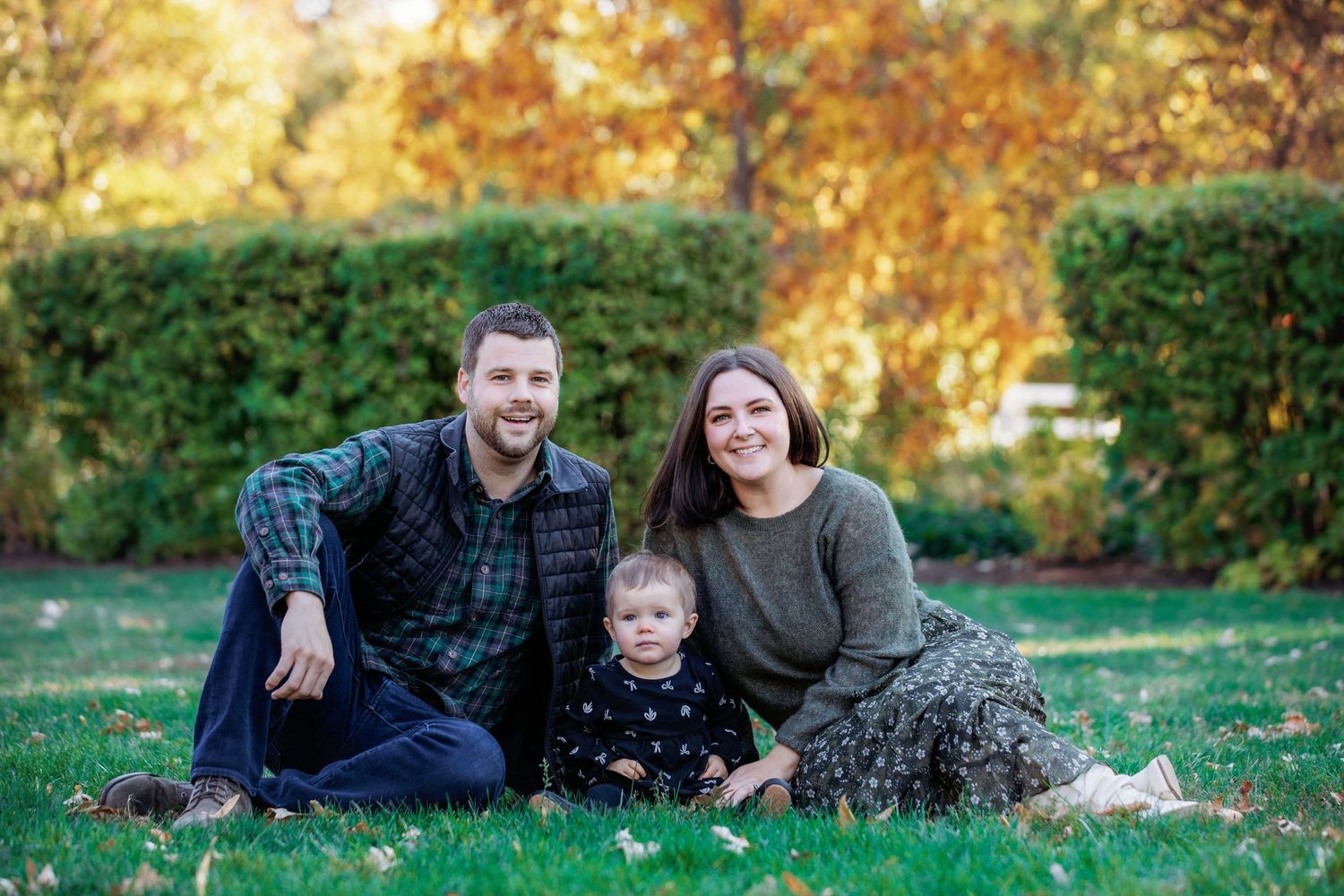 A family sits together on grass during autumn with colorful fall foliage and green hedges in the background.