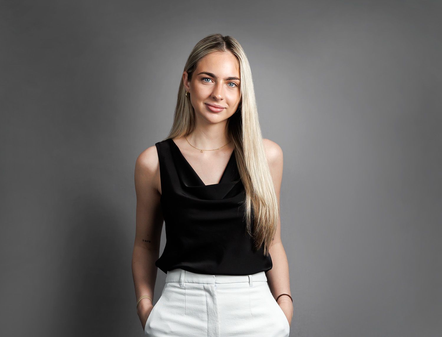 Modern corporate portrait of a young woman with long blonde hair wearing black sleeveless top and white pants against a gray background
