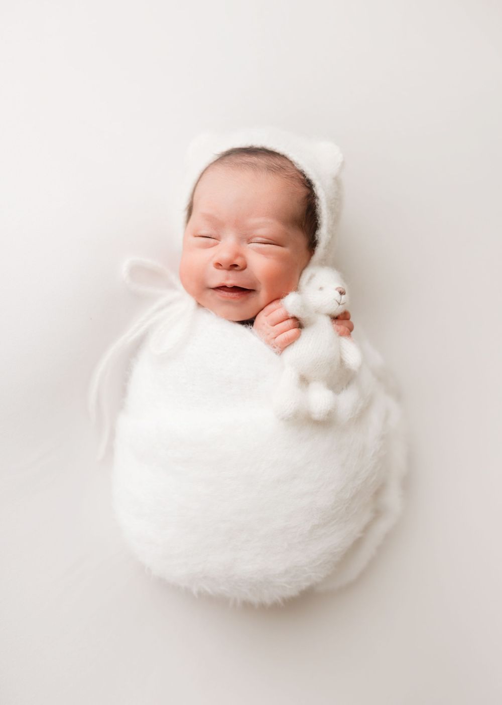 Newborn baby wrapped in soft white blanket smiling while holding small plush toy.