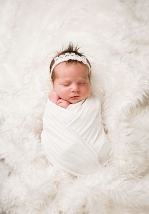 A swaddled newborn with a lace headband rests on soft white fur.