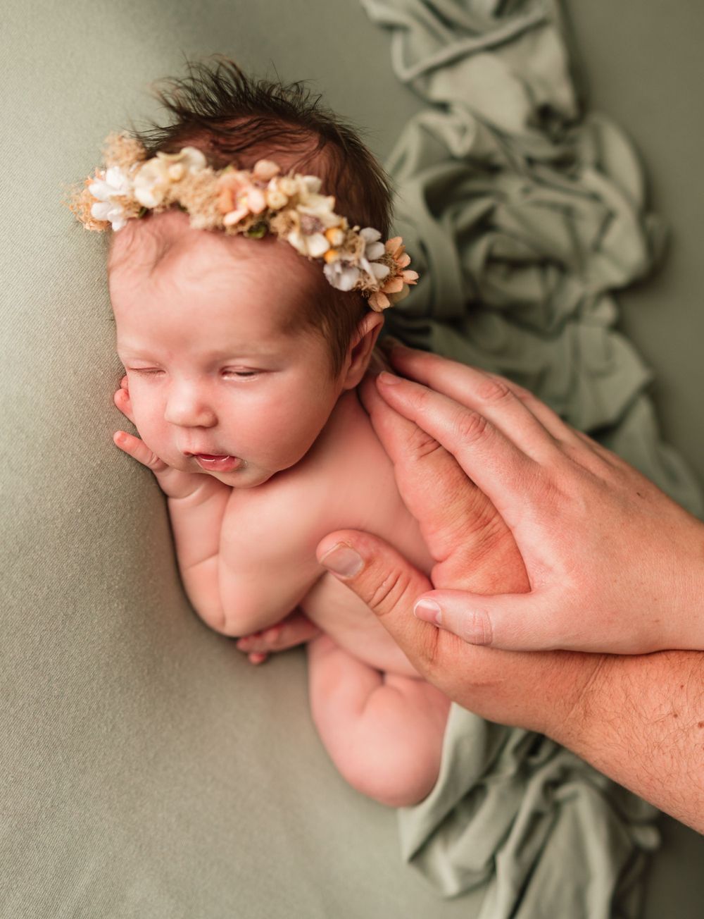 A sleeping newborn with floral crown is gently supported by caring hands against sage green fabric.