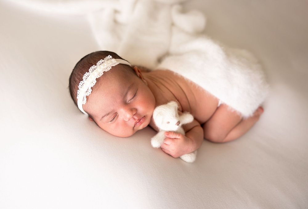 Sleeping newborn with lace headband holding small plush toy on white background.
