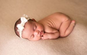 A peaceful newborn wearing a delicate lace headband rests on a beige blanket.