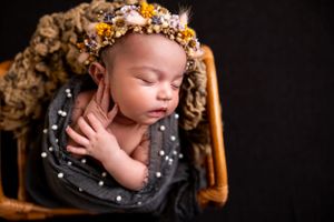 A sleeping newborn wearing a floral crown lies peacefully in a wooden chair against a dark background.