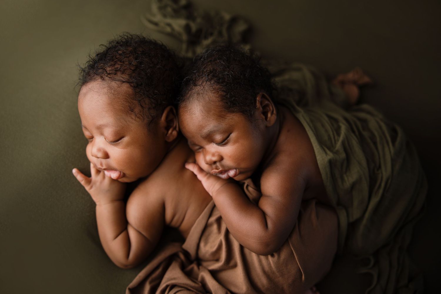 Two sleeping newborn babies lying side by side on a brown blanket against dark green backdrop. Arkansas Newborn Photographer