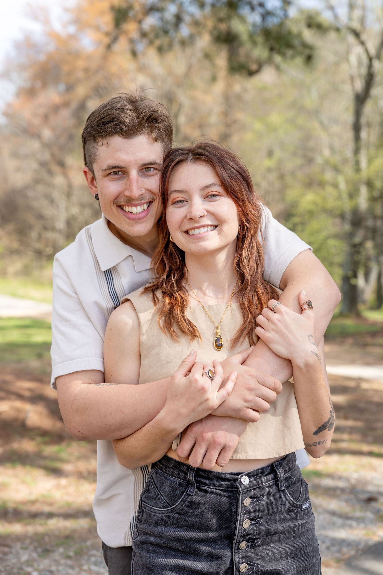 A joyful couple embraces while wearing casual attire outdoors with trees in the background on a sunny day.