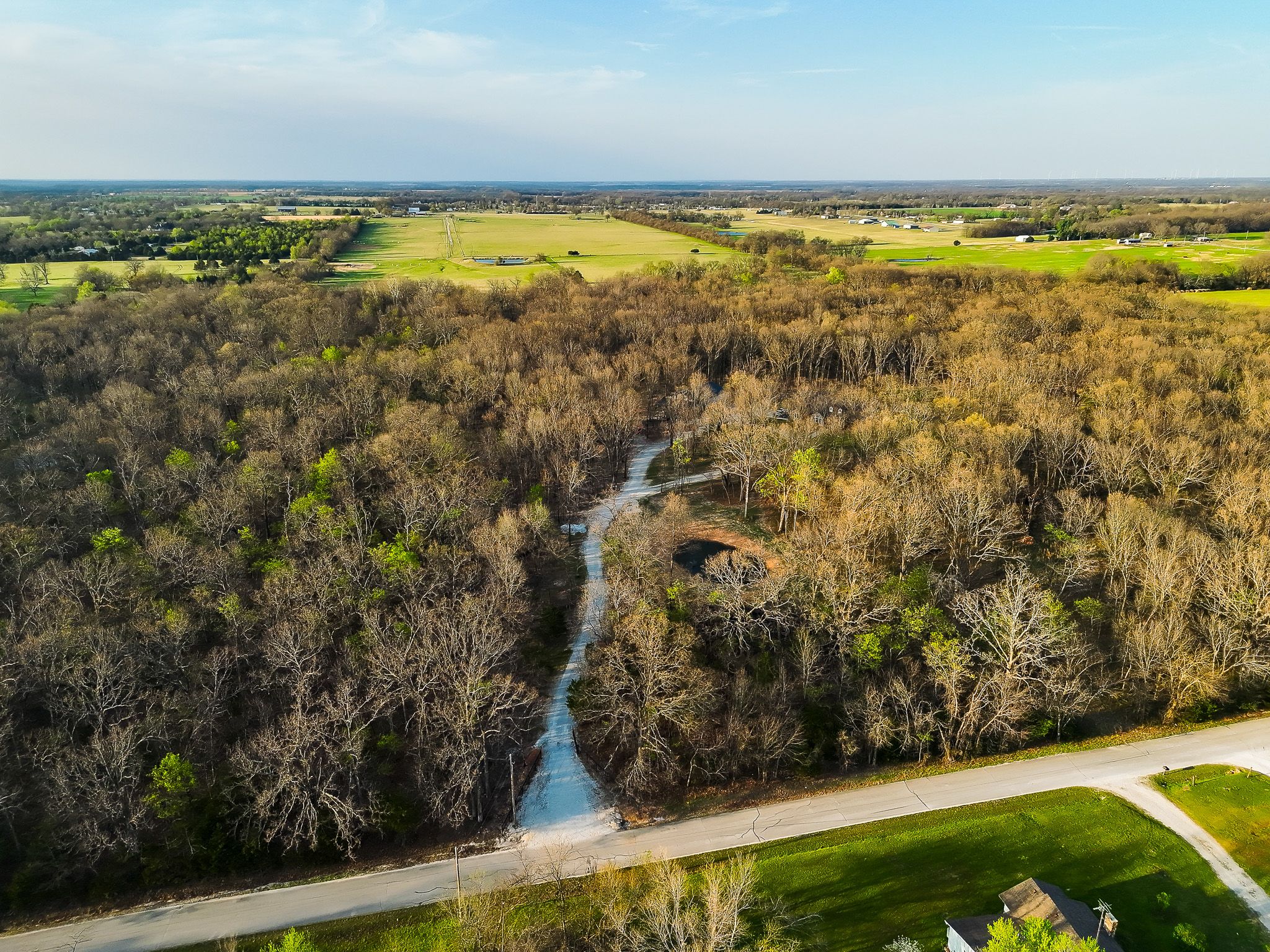 Aerial view of a winding creek through dense woodland with open farmland and blue skies in the background.