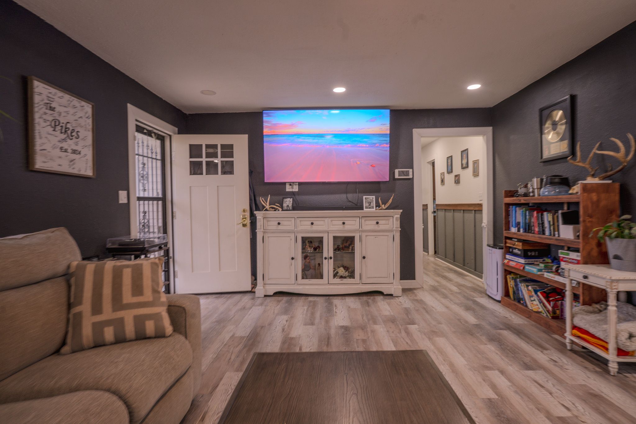 Cozy living room with gray walls, hardwood floors, wall-mounted TV, white cabinet, and bookshelf with colorful books.