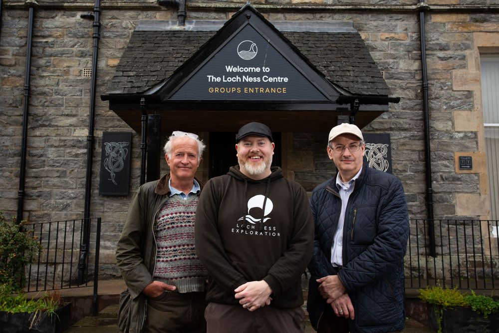 Three men pose outside the Loch Ness Centre groups entrance in Scotland.