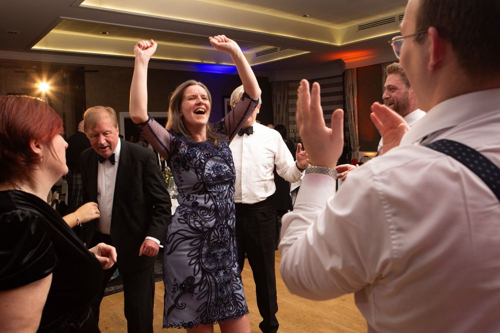 Woman dancing joyfully with arms raised at a lively party, surrounded by smiling guests in elegant attire.