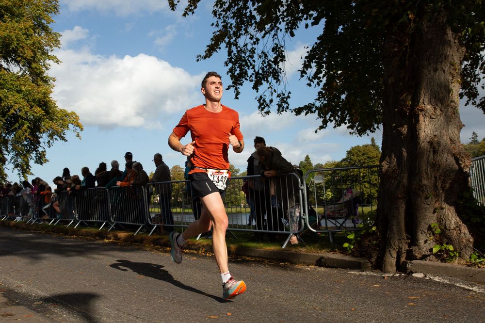 Man in red shirt running a road race past metal barriers and spectators on a sunny day.