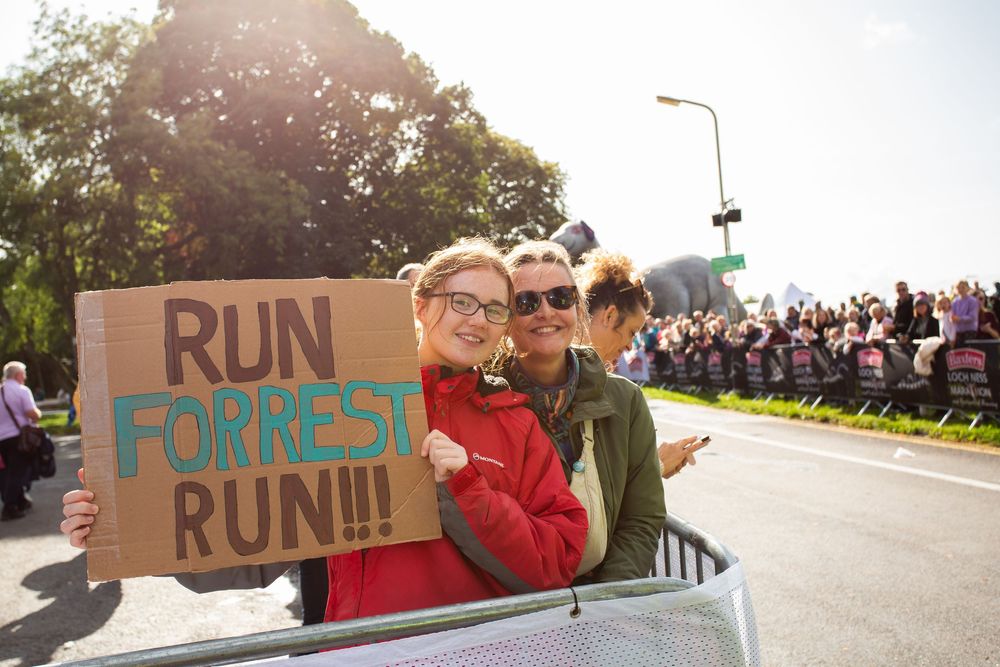 Two women smile holding a "Run Forrest Run" sign at a road race event with crowds cheering in the background.