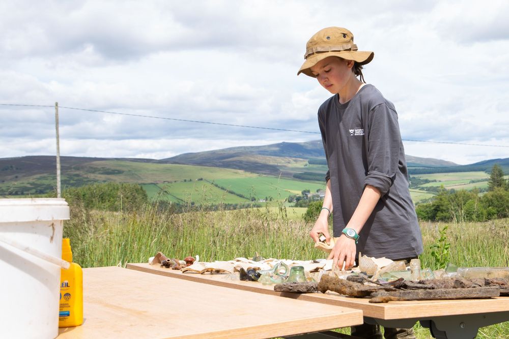 Young archaeologist examines artifacts on wooden table at outdoor dig site with scenic green hills in background.