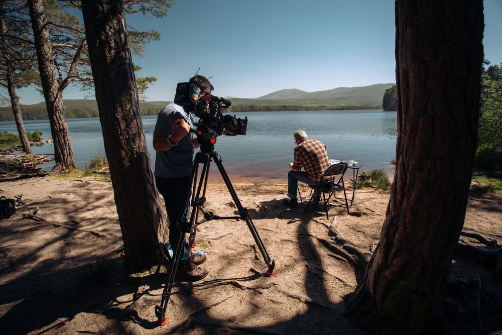 Filmmaker operating a professional camera on tripod by a scenic lake shore, with a person sitting nearby among pine trees.