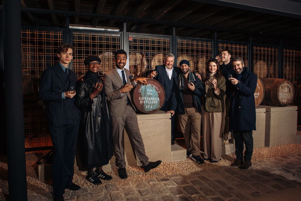 Group of people posing together near wooden barrels in a winery, smiling and holding drinks.