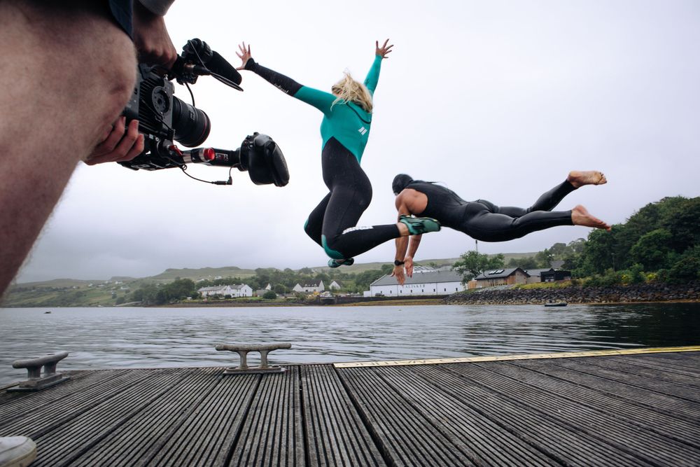 Two people leap off a wooden dock into a lake while a cameraman films them on a cloudy day.
