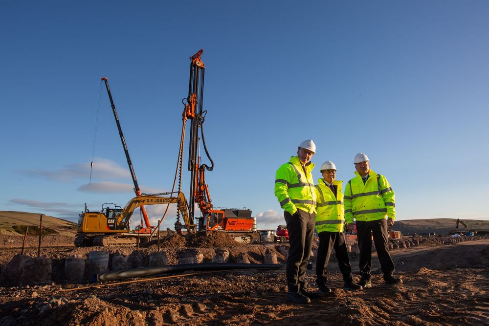 Three workers in hi-vis jackets and hard hats stand at a construction site with a drilling rig under a clear blue sky.