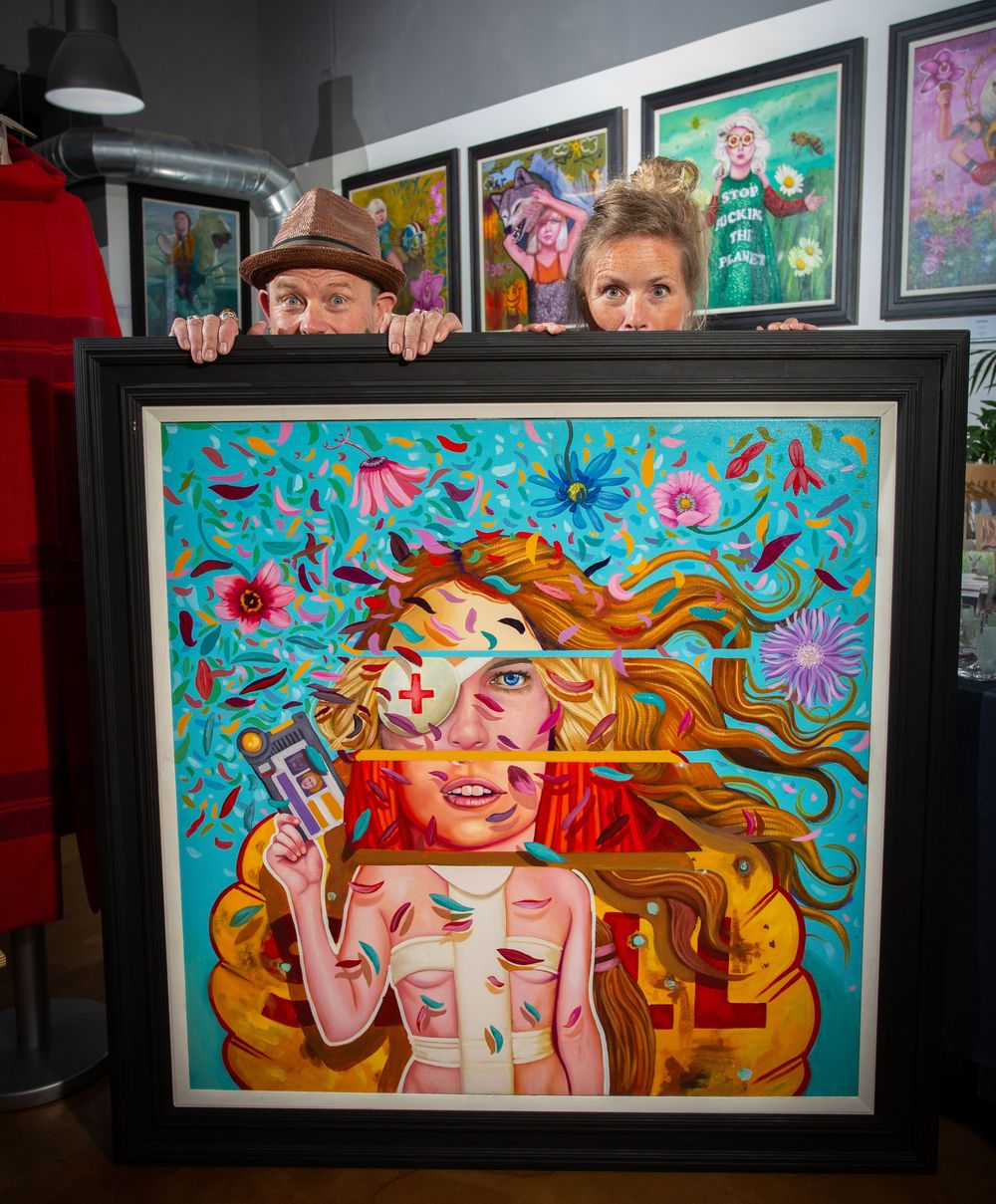 Two artists peek over a vibrant framed painting of a woman with flowing red hair and colorful flowers in a gallery.