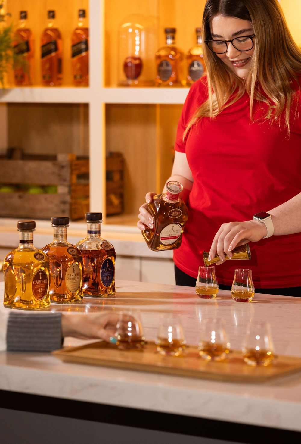Woman in red shirt pouring whiskey samples at a tasting bar with Crown Royal bottles on display.