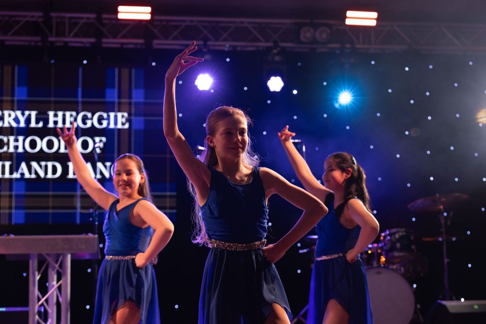 Three girls in blue dresses perform on stage at Cheryl Heggie School of Highland Dancing recital.