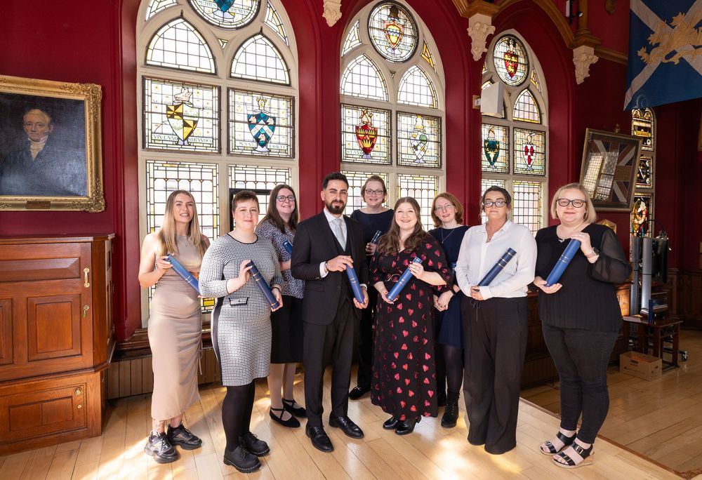 Group of professionals posing together in an ornate hall with stained glass windows and heraldic crests.