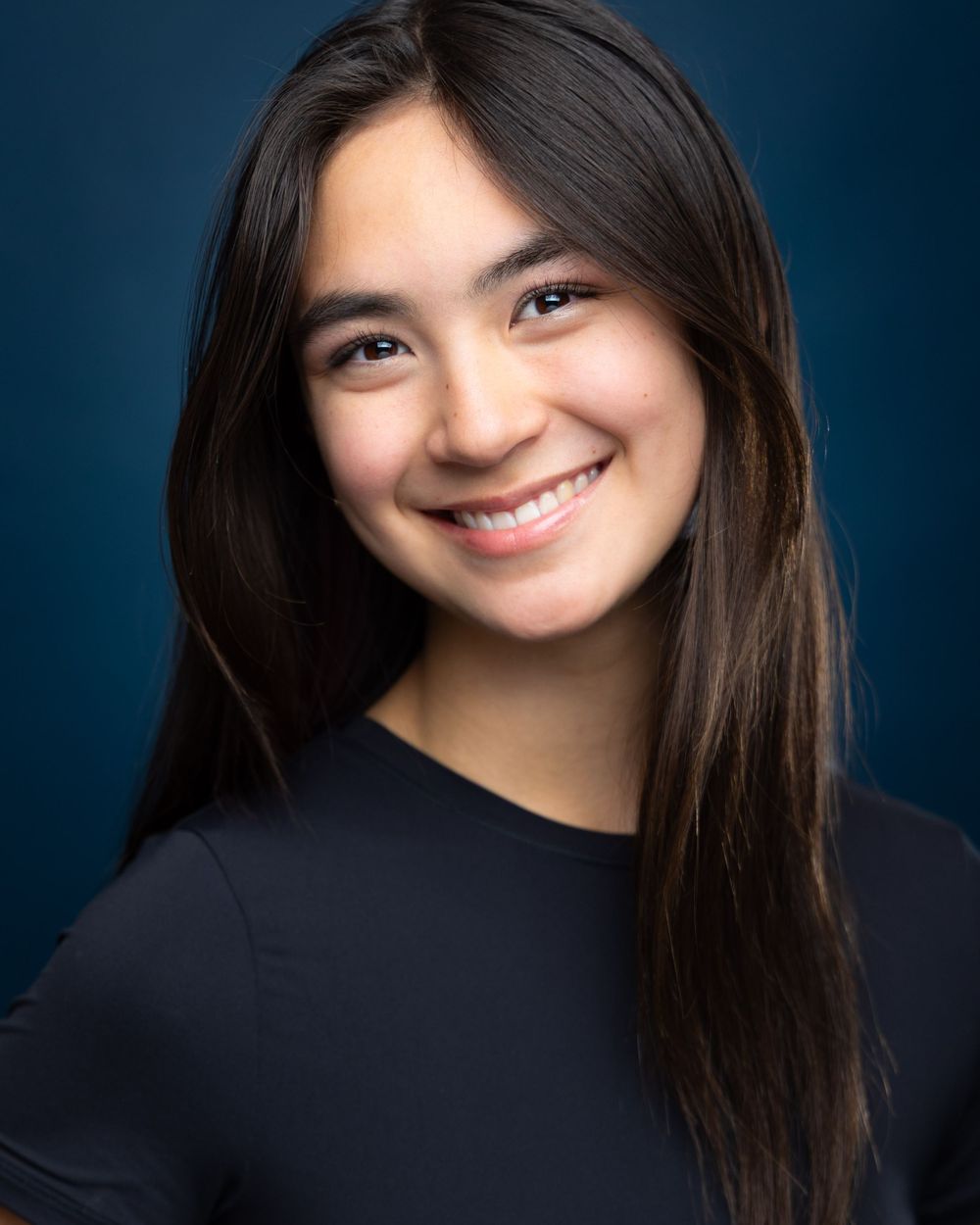 Young woman with long dark hair smiles confidently in a professional headshot against a dark blue background.