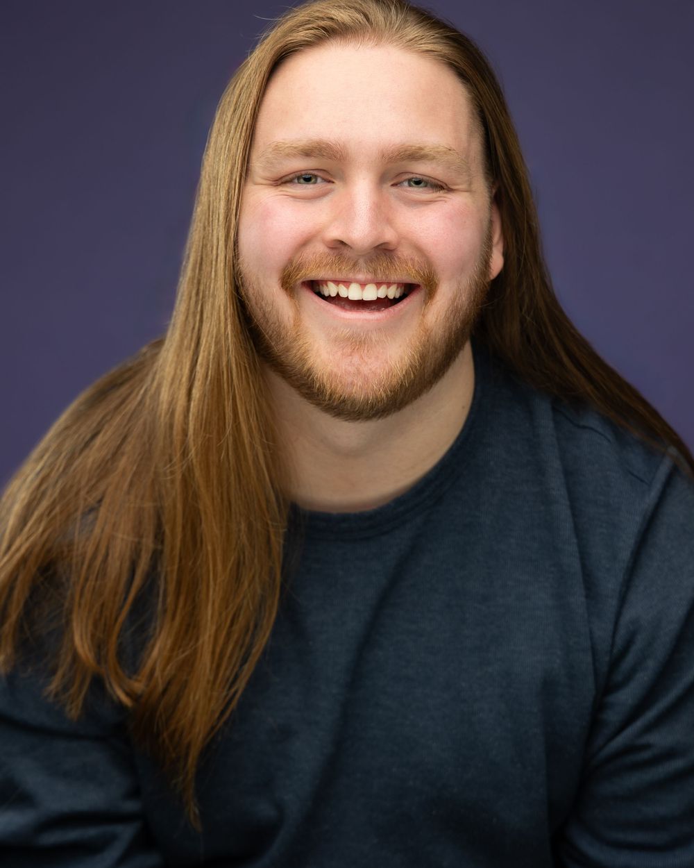 Portrait showing someone with long hair and beard smiling against a purple background.