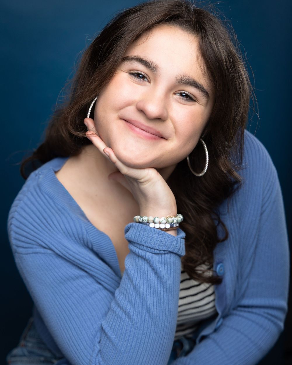 Smiling teenage girl with hoop earrings and beaded bracelet wearing a blue sweater poses against a dark blue background.