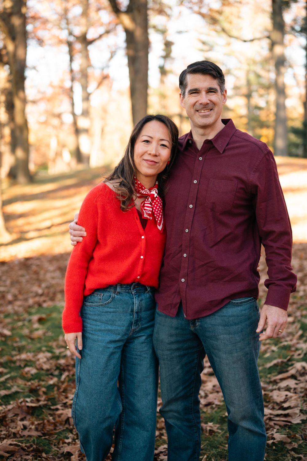 A couple in matching burgundy and red outfits stands together on fallen autumn leaves.