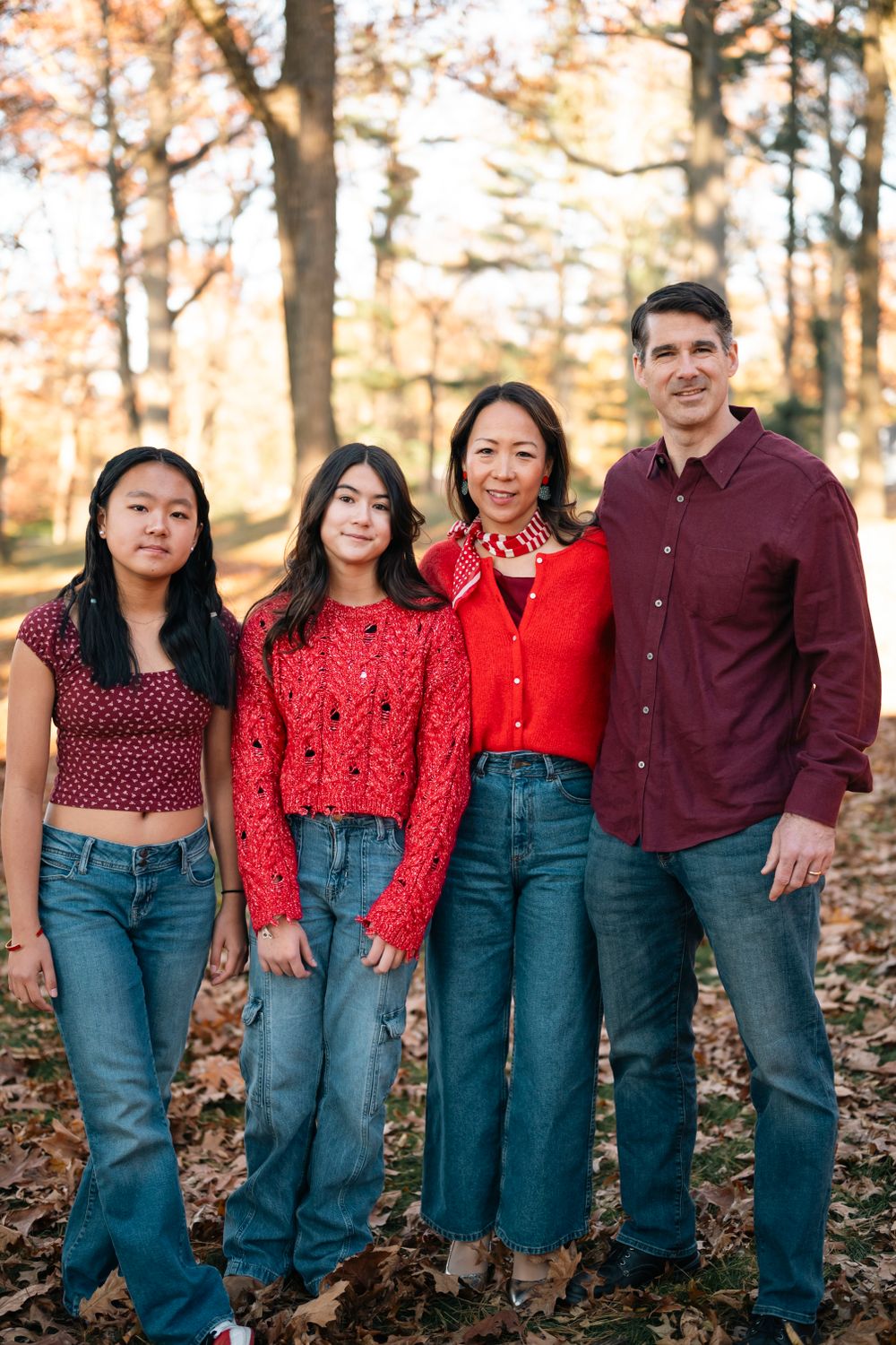 A family of four wearing coordinated casual outfits poses together in a fall forest setting.