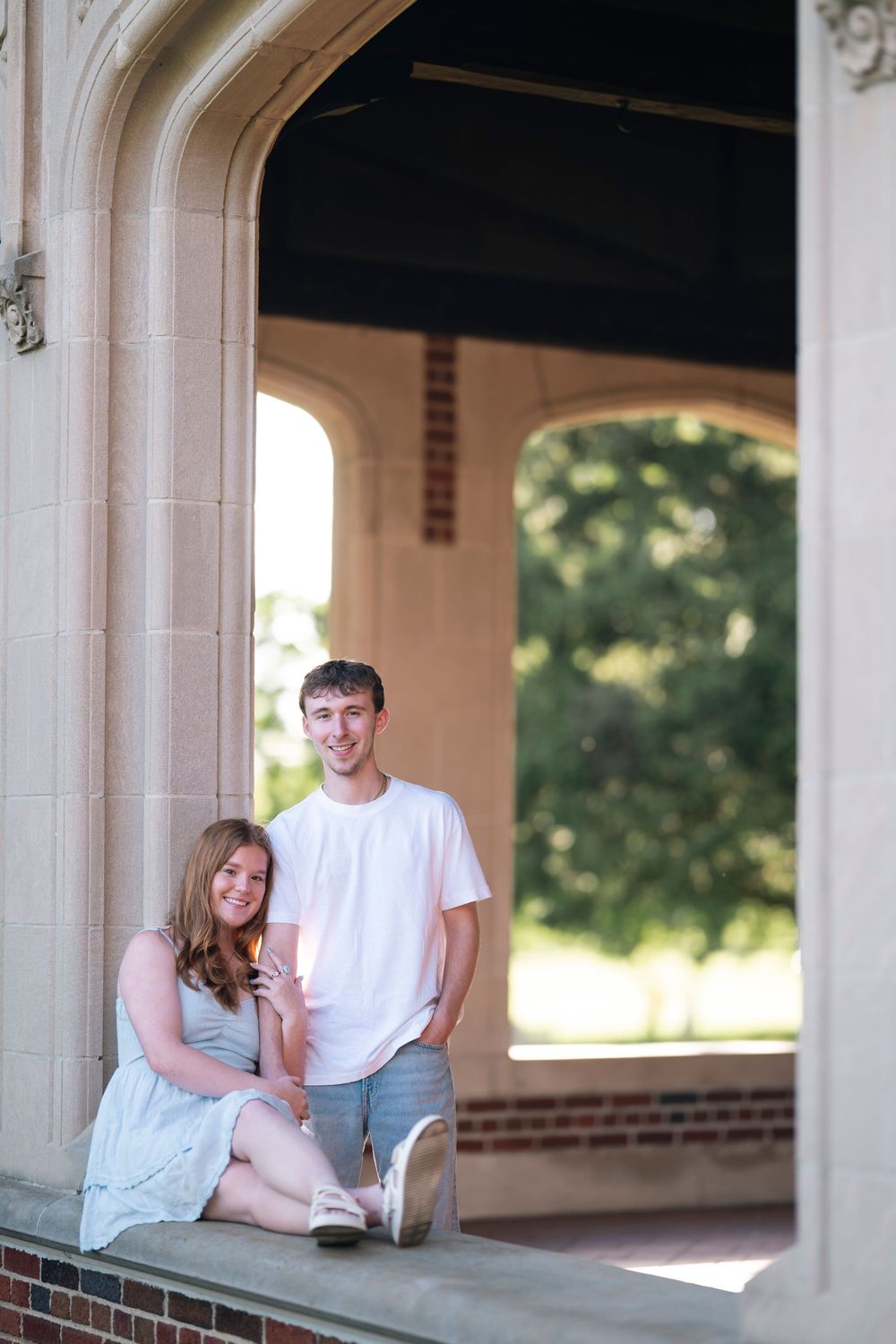 A couple poses together under a stone archway on a bright summer day at a brick building.
