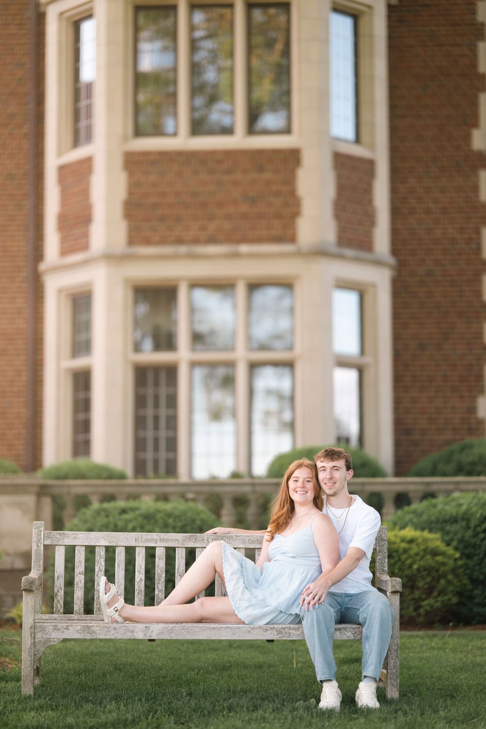 Young couple relaxes on wooden bench outside brick building with large windows on sunny afternoon.