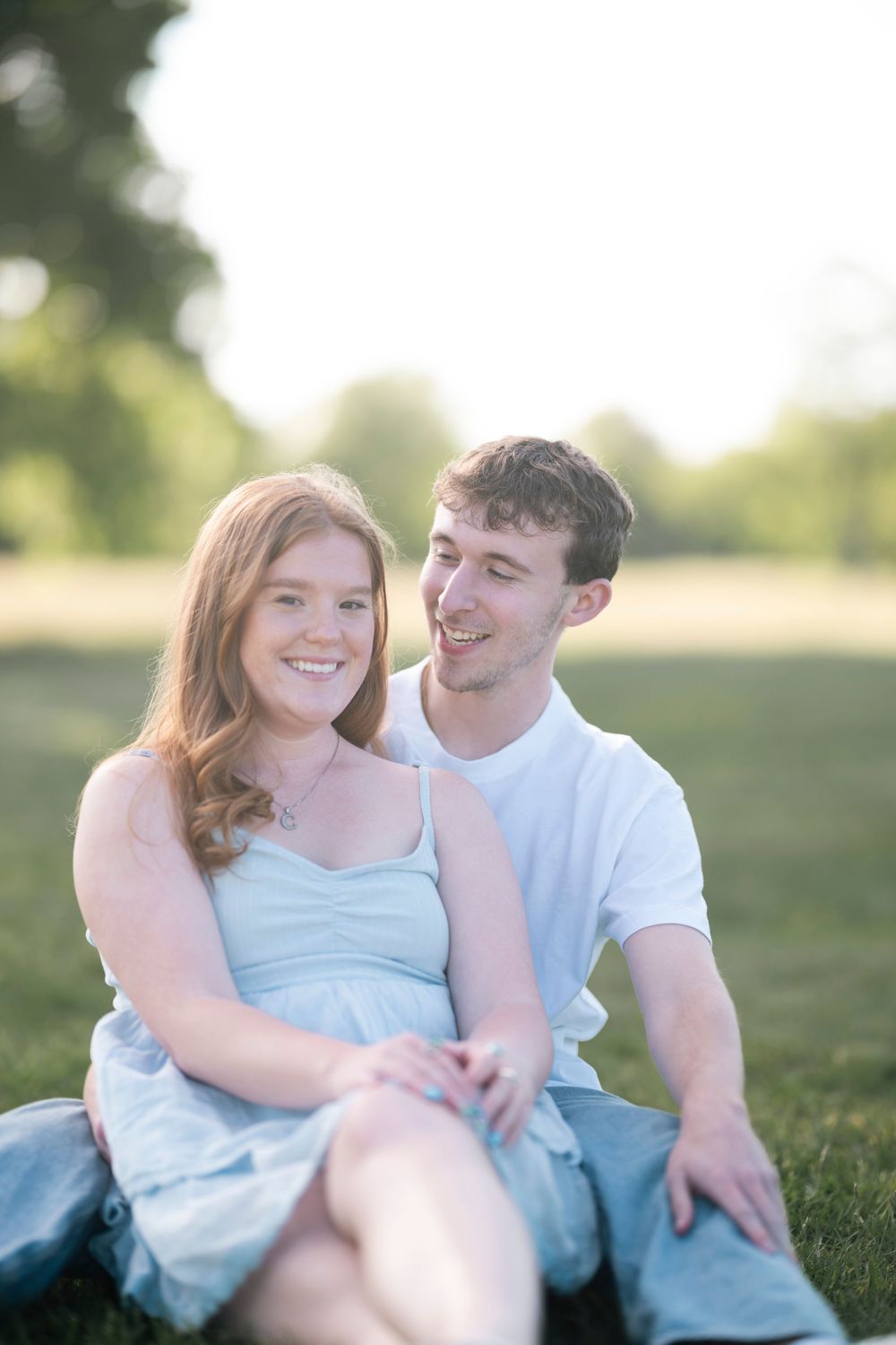 A couple sits together on grass in a park during golden hour wearing coordinating light blue and white outfits.